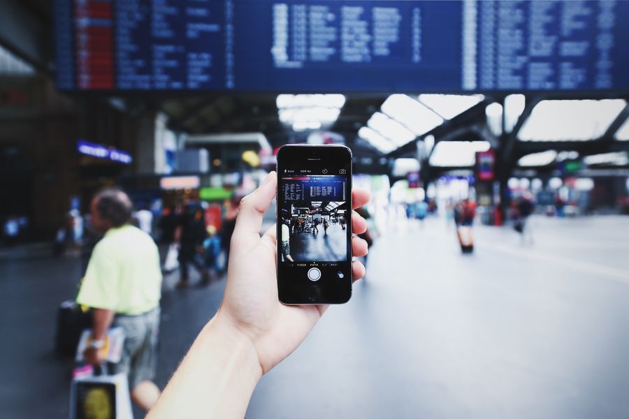 A hand holding a smartphone capturing a bustling train station with blurred travelers in the background. — Marchés nocturnes et trains tardifs — connecté tout du long.