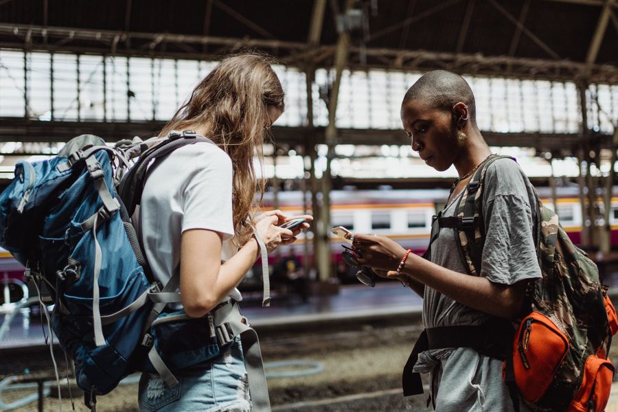 Two adventurous backpackers using smartphones while waiting at a bustling train station. — La longue escale : streaming, travail et messages, tout couvert.